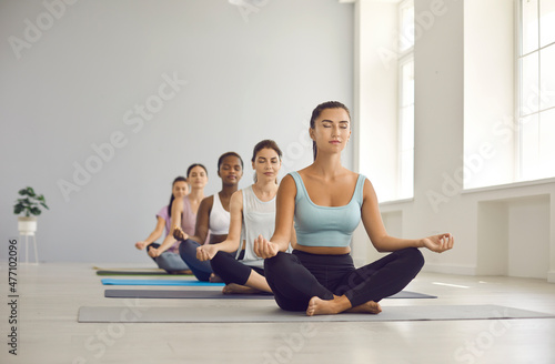 Group of young sports different women practicing yoga while sitting in Suhasan pose. Women with closed eyes sit in row one after the other and meditate together in bright yoga studio.