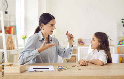 Female speech therapist working on dyslexic kid's problems and impediments. Happy child together with speech language pathologist sitting at a desk in modern office, playing fun game, learning letters
