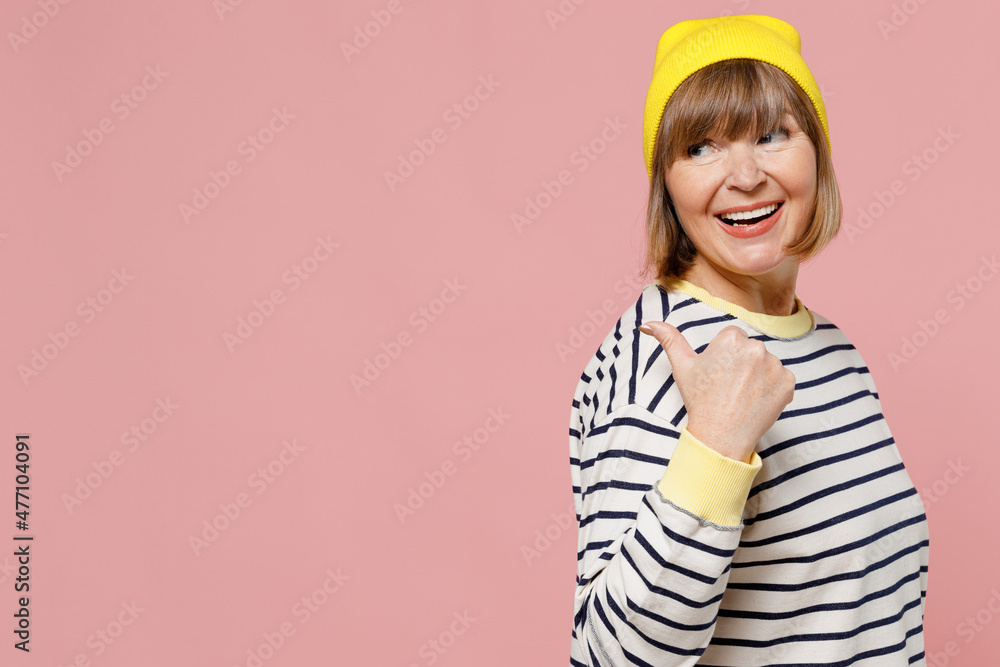 Elderly stylish woman 50s in striped shirt yellow hat point thumb finger aside on workspace area mock up area isolated on plain pastel light pink background studio portrait. People lifestyle concept. Elderly stylish woman 50s in striped shirt yellow hat point thumb finger aside on workspace area mock up area isolated on plain pastel light pink background studio portrait. People lifestyle concept.