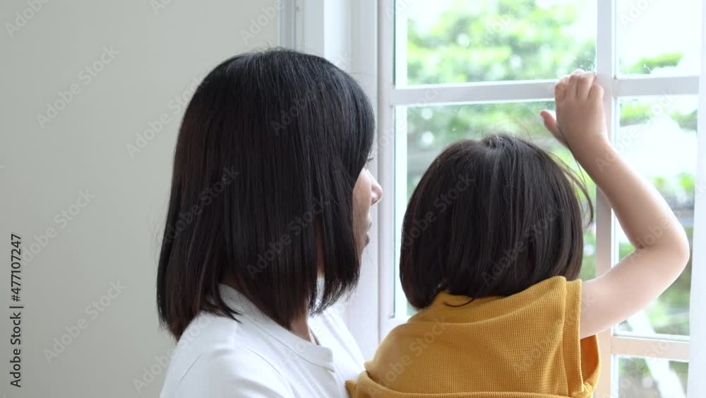 An Asian mother holds her son looking through a window with white curtains. Cute son smiles shyly and looks at the camera. Happy family. An Asian mother holds her son looking through a window with white curtains. Cute son smiles shyly and looks at the camera. Happy family.