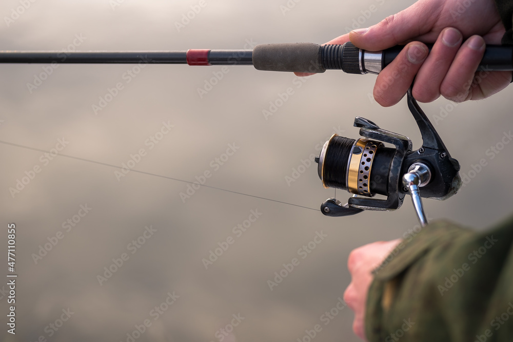 The frozen hands of a male angler close-up to hold the spinning motion wind the fishing line on a reel against the background of a winter river. Selective focus on the reel. Background The frozen hands of a male angler close-up to hold the spinning motion wind the fishing line on a reel against the background of a winter river. Selective focus on the reel. Background