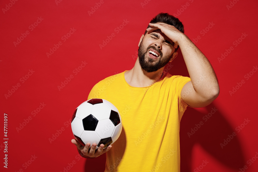Sick ill tired young bearded man football fan in yellow t-shirt cheer up support favorite team hold soccer ball hand on forehead feel bad seedy isolated on plain dark red background studio portrait. Sick ill tired young bearded man football fan in yellow t-shirt cheer up support favorite team hold soccer ball hand on forehead feel bad seedy isolated on plain dark red background studio portrait.