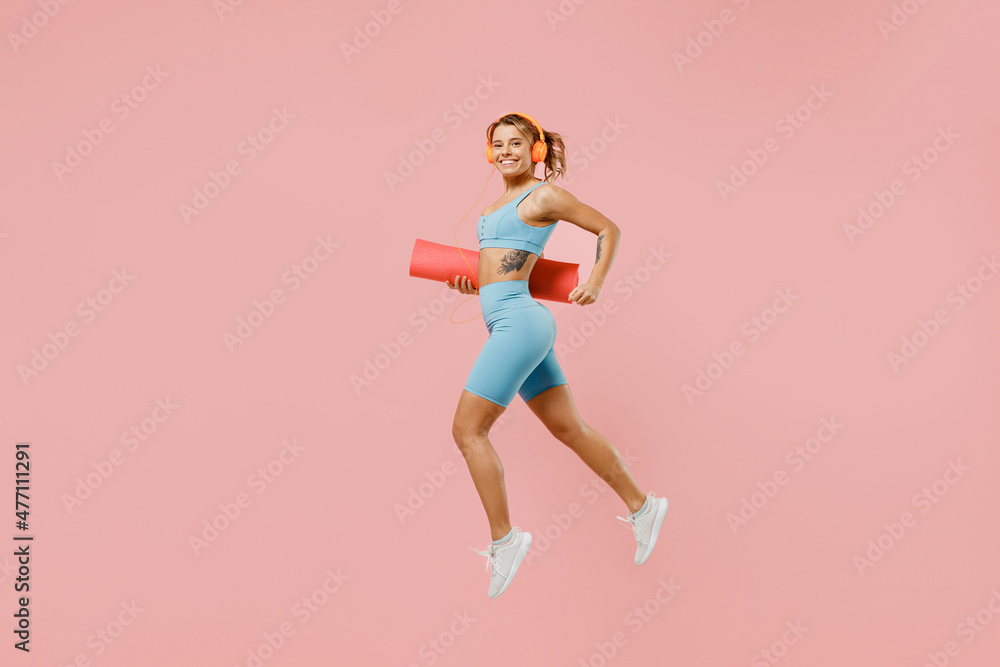 Full body young strong sporty fitness trainer instructor woman wear blue tracksuit headphones spend time in home gym hold mat jump high isolated on pastel plain pink background. Workout sport concept.