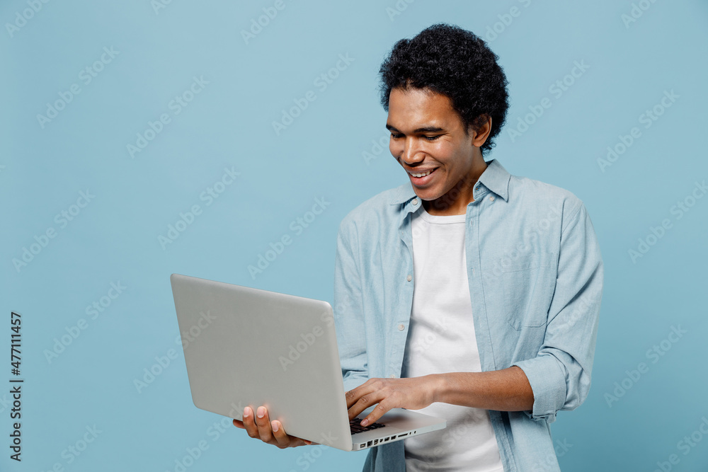 Smiling vivid young black curly man 20s years old wear white shirt hold use work on laptop pc computer typing browsing chatting send sms isolated on plain pastel light blue background studio portrait Smiling vivid young black curly man 20s years old wear white shirt hold use work on laptop pc computer typing browsing chatting send sms isolated on plain pastel light blue background studio portrait