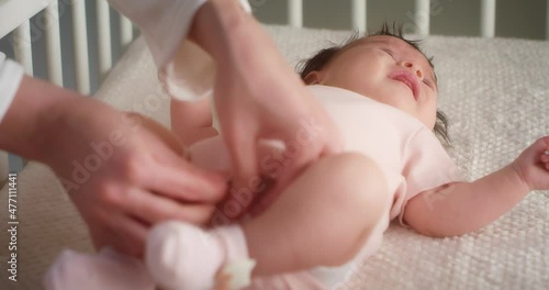 Mom lovingly puts a pink bodysuit on a newborn baby girl lying in a diaper on her back in a child crib. Authentic shots of a neonate toddler in the nursery.The concept of childhood,New life,Fatherhood