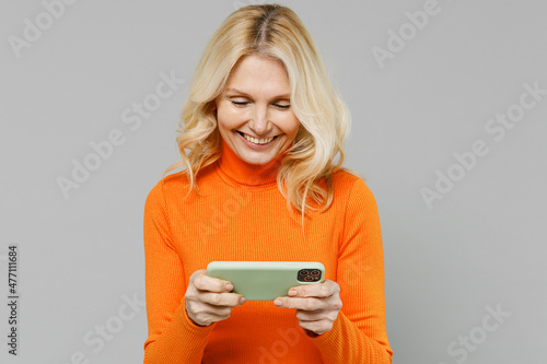 Gambling elderly smiling cool happy blonde woman 50s in orange turtleneck using play racing app on mobile cell phone hold gadget smartphone for pc video games isolated on plain grey background studio.