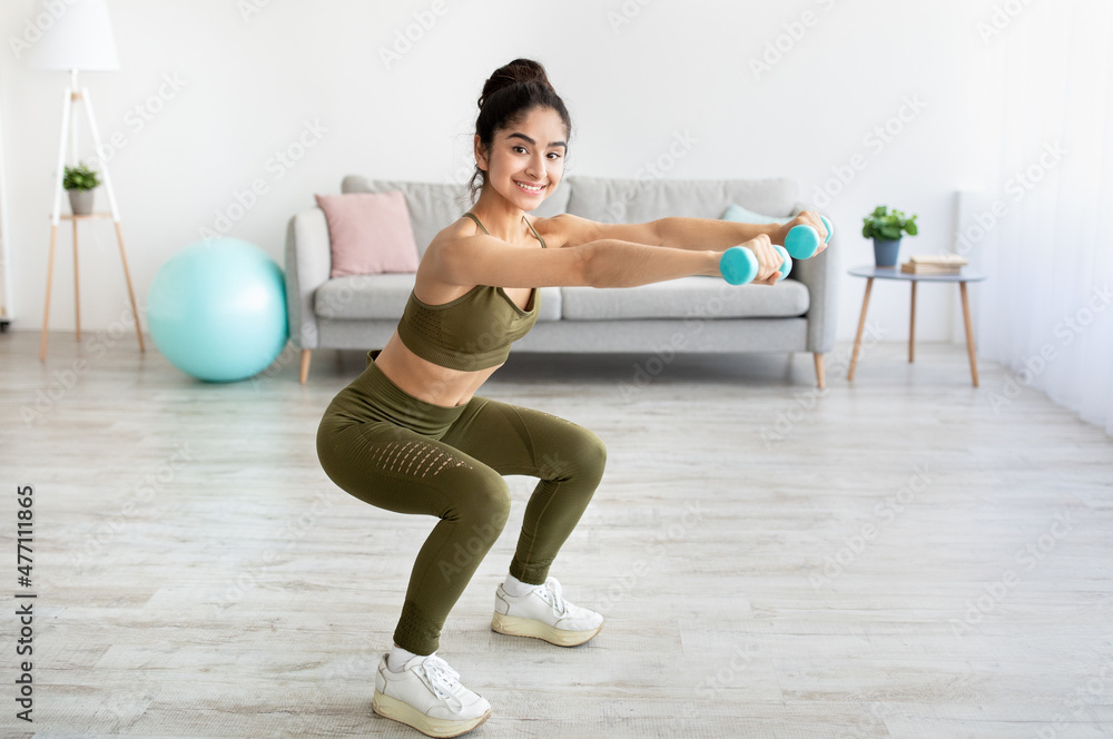 Strong young Indian woman working out with dumbbells, squatting at home, empty space Strong young Indian woman working out with dumbbells, squatting at home, empty space