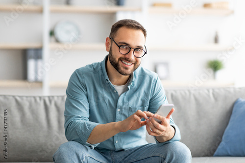 Cheerful millennial guy in glasses sitting on cozy couch with cellphone and messaging to his friend, indoors © Prostock-studio