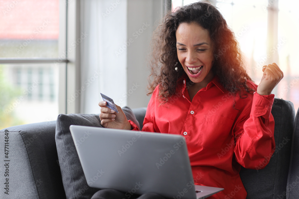 Happy woman sitting and using computer and holding credit card for online payment or shopping in living room. Businesswoman showing payment completed with raising hands and exciting emotion. Happy woman sitting and using computer and holding credit card for online payment or shopping in living room. Businesswoman showing payment completed with raising hands and exciting emotion.