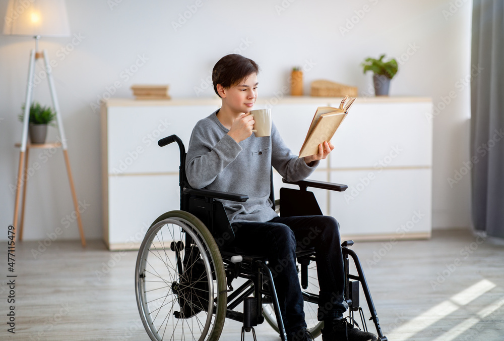 Positive handicapped teen boy in wheelchair reading interesting book and drinking coffee at home Positive handicapped teen boy in wheelchair reading interesting book and drinking coffee at home