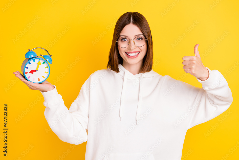 Photo of adorable sweet young woman dressed white sweatshirt showing thumb up holding timer smiling isolated yellow color background Photo of adorable sweet young woman dressed white sweatshirt showing thumb up holding timer smiling isolated yellow color background