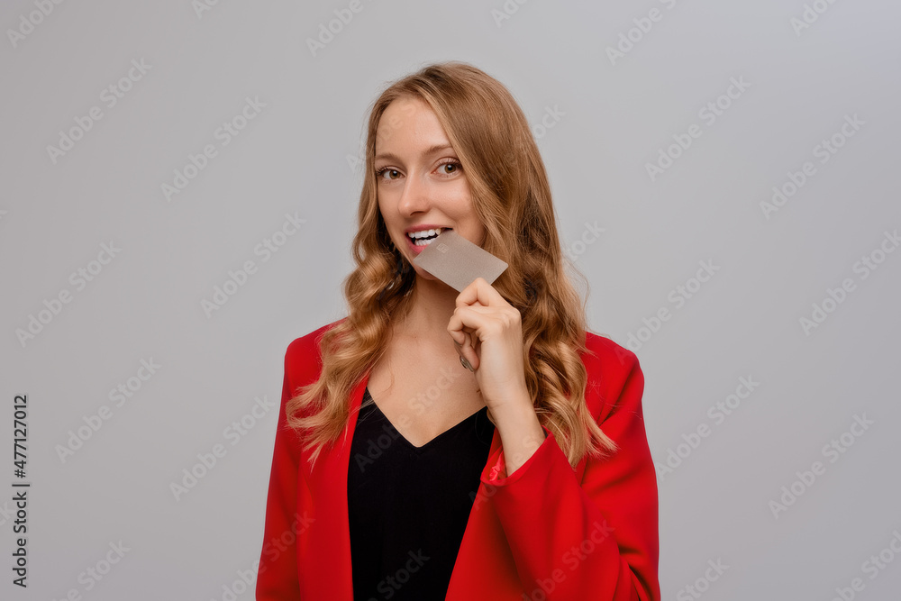 Playful young blonde woman biting credit card, thinking of doing online shopping, standing in red blazer against gray background Playful young blonde woman biting credit card, thinking of doing online shopping, standing in red blazer against gray background