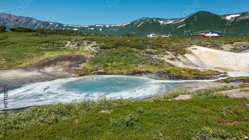 Blue thermal lake in the caldera of the extinct volcano Uzon. Sulphurous deposits on the soil, green vegetation around. Helicopters parked in the distance. Mountains against the sky. Kamchatka