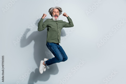 Photo of lucky retired man wear green shirt glasses rising fists jumping high empty space isolated grey color background