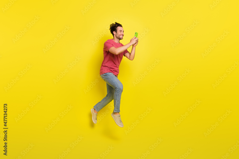 Full size photo of nice beard millennial guy jump look telephone wear red t-shirt jeans sneakers isolated on yellow background