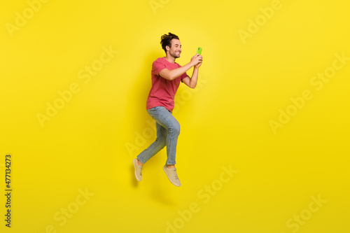 Full size photo of nice beard millennial guy jump look telephone wear red t-shirt jeans sneakers isolated on yellow background