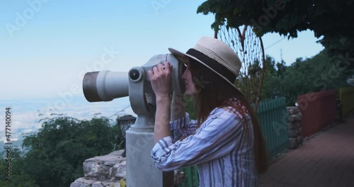 Happy woman traveler looking through viewing binoculars at Tirana city panorama. Caucasian woman in straw hat on viewpoint watching in telescope of sights observes summer cityscape view Daitit Albania