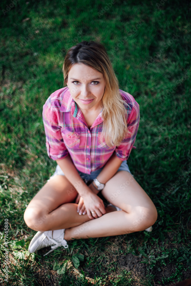 Photo from above of adult shining blonde woman sitting on grass in park wearing checkered shirt, jeans skirt and white sneakers in daytime. Adorable background full of green. Photo from above of adult shining blonde woman sitting on grass in park wearing checkered shirt, jeans skirt and white sneakers in daytime. Adorable background full of green.