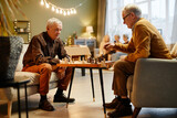 Two senior Caucasian men sitting on armchairs in front of each other in living room and playing chess while three aged women sitting on background