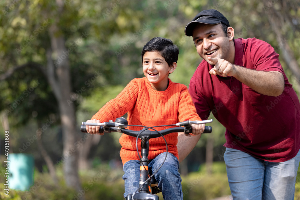 Cheerful father teaching son riding bicycle while admiring view at park.
Cheerful father teaching son riding bicycle while admiring view at park.