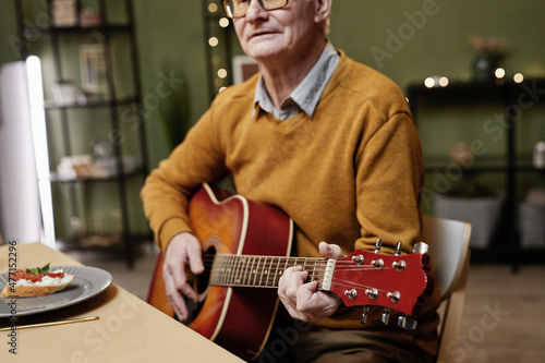 Senior Caucasian man wearing casual clothes and eyeglasses sitting at dining table and playing acoustic guitar