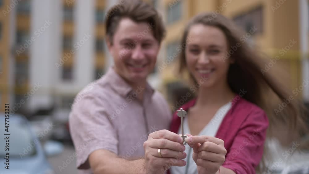 Close-up key in couple hands with blurred Caucasian man and woman talking smiling at background. Happy husband and wife buying new apartment posing outdoors in slow motion Close-up key in couple hands with blurred Caucasian man and woman talking smiling at background. Happy husband and wife buying new apartment posing outdoors in slow motion