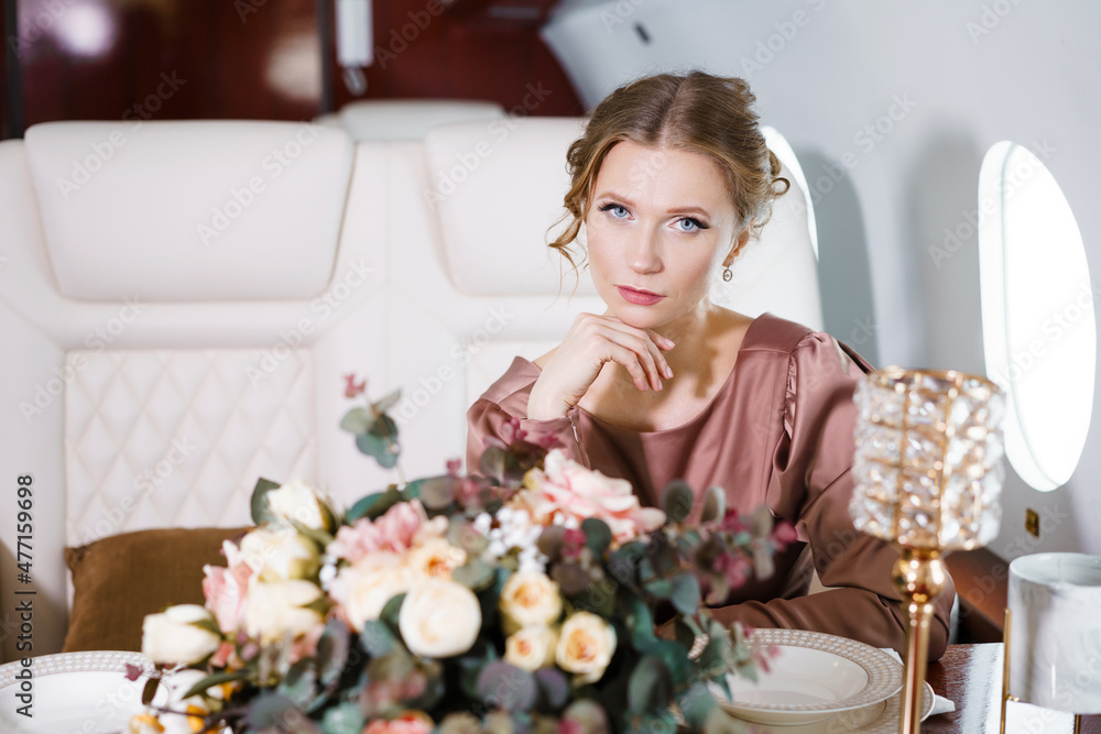 Beautiful woman in private jet in stylish dress with hairstyle and make-up sits at festive table with a bouquet of flowers. In a passenger seat in a comfortable plane by the window. Beautiful woman in private jet in stylish dress with hairstyle and make-up sits at festive table with a bouquet of flowers. In a passenger seat in a comfortable plane by the window.