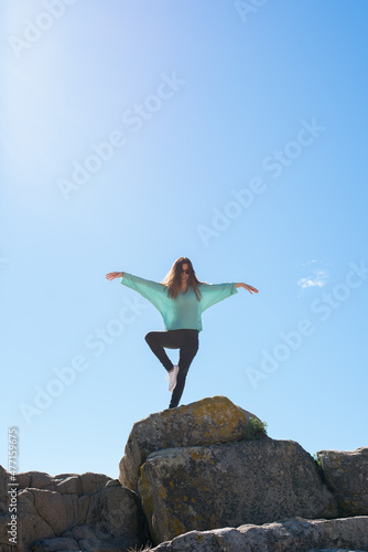 Young happy woman with sunglasses stands on a rock and tries to raise her hands up in a yoga pose during summer vacation. Relaxation and meditation yoga concept.