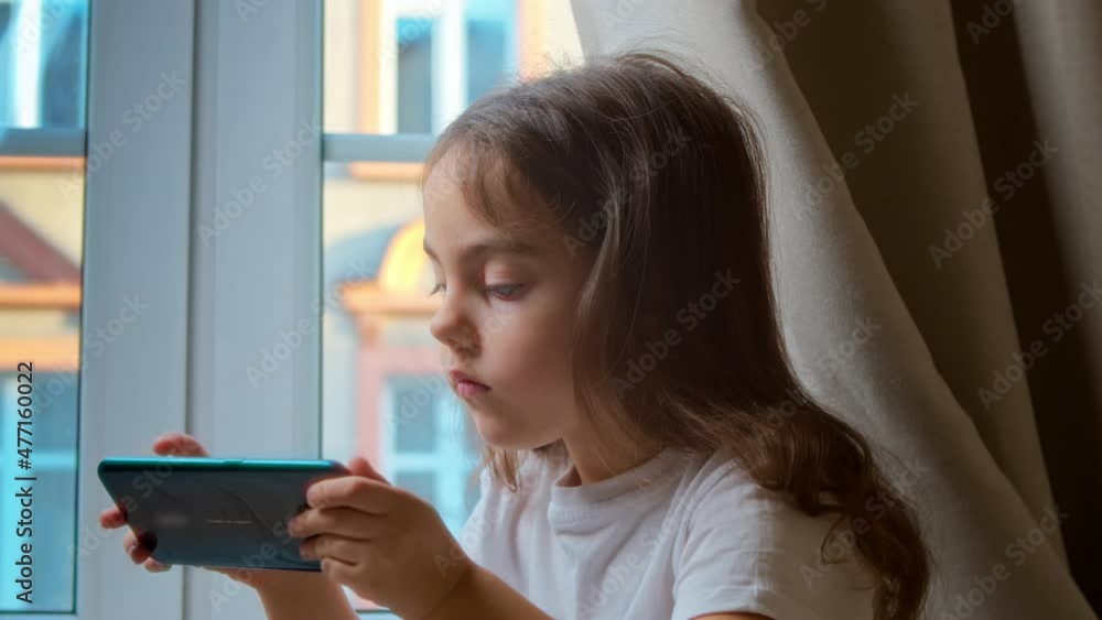 little preschool girl is sitting on the bed in the bedroom by the window, using a smartphone, playing video games, watching cartoons, spending time on weekends with a modern technological concept. little preschool girl is sitting on the bed in the bedroom by the window, using a smartphone, playing video games, watching cartoons, spending time on weekends with a modern technological concept.