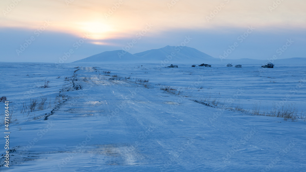 A road among the snow-covered tundra in the Arctic. View of the road, old abandoned houses in the tundra and snow-capped mountains. Winter Arctic landscape. Travel to the Far North of Russia. Chukotka A road among the snow-covered tundra in the Arctic. View of the road, old abandoned houses in the tundra and snow-capped mountains. Winter Arctic landscape. Travel to the Far North of Russia. Chukotka