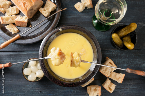 Swiss cheese fondue as new year party meal with bread on long forks, pickles and wine on a dark wooden table, high angle view from above, selected focus