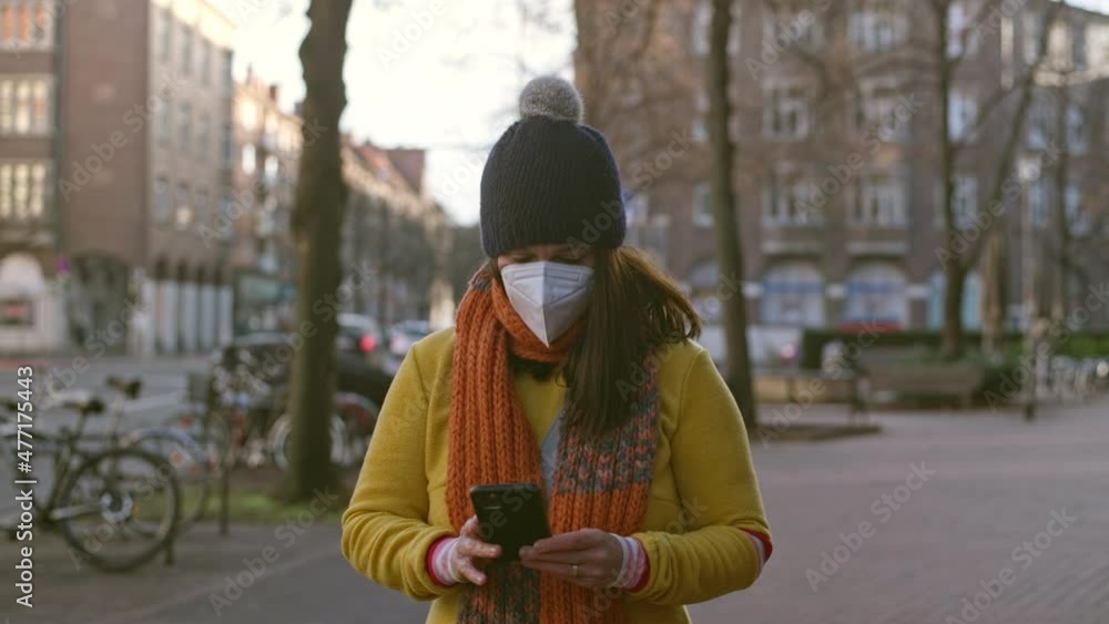 Middle-aged brunette woman wearing a face mask walking on a sidewalk in winter while typing on her mobile phone, concept of technology and health care Middle-aged brunette woman wearing a face mask walking on a sidewalk in winter while typing on her mobile phone, concept of technology and health care