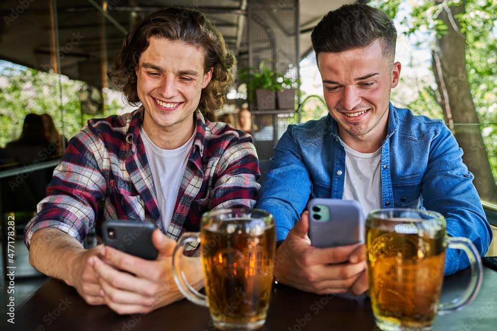Two friends using mobile phone and drinking beer in pub.