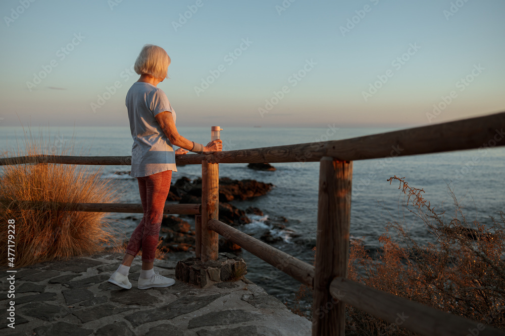 Rear view of aged woman in sportswear, enjoying beauty of nature while looking at sea coast in evening sunset