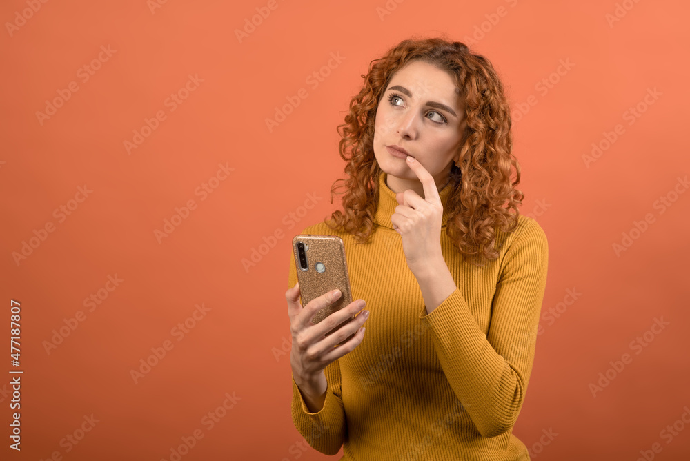 Young and attractive redhead Caucasian girl in orange jumper using smartphone with a pensive look isolated on orange studio background. Young and attractive redhead Caucasian girl in orange jumper using smartphone with a pensive look isolated on orange studio background.