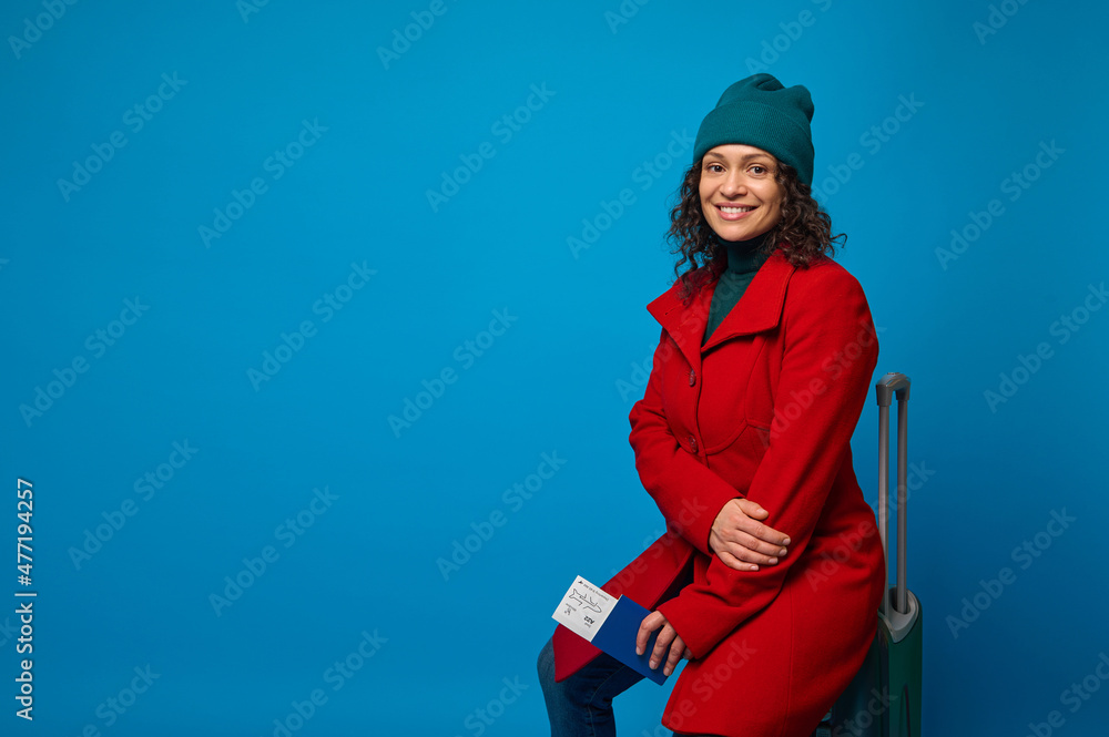Delightful woman in red coat, green hat and pullover, with passport, air ticket, boarding pass, sits on suitcase and smiles toothy smile looking at camera. Travel. Tourism concept. Blue background