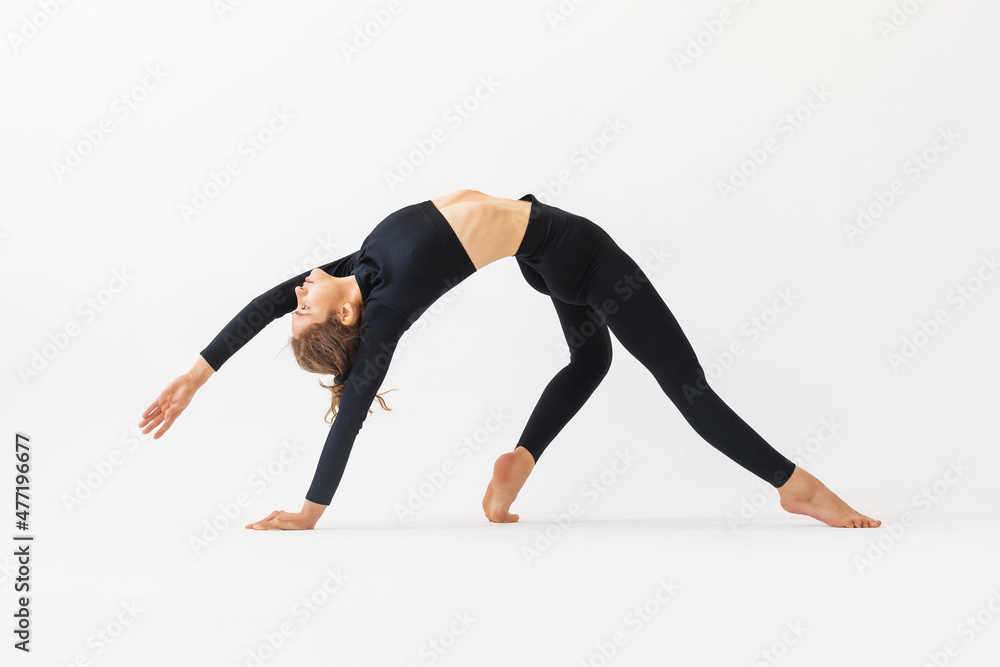 A young woman practicing yoga performs the exercise of Kamatkarasana, the pose of a dancing dog, trains in black sportswear on a white background