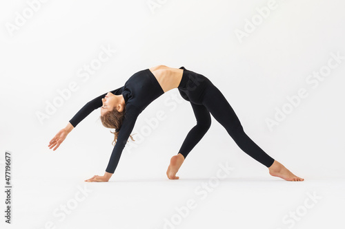 A young woman practicing yoga performs the exercise of Kamatkarasana, the pose of a dancing dog, trains in black sportswear on a white background © Sergey Chayko