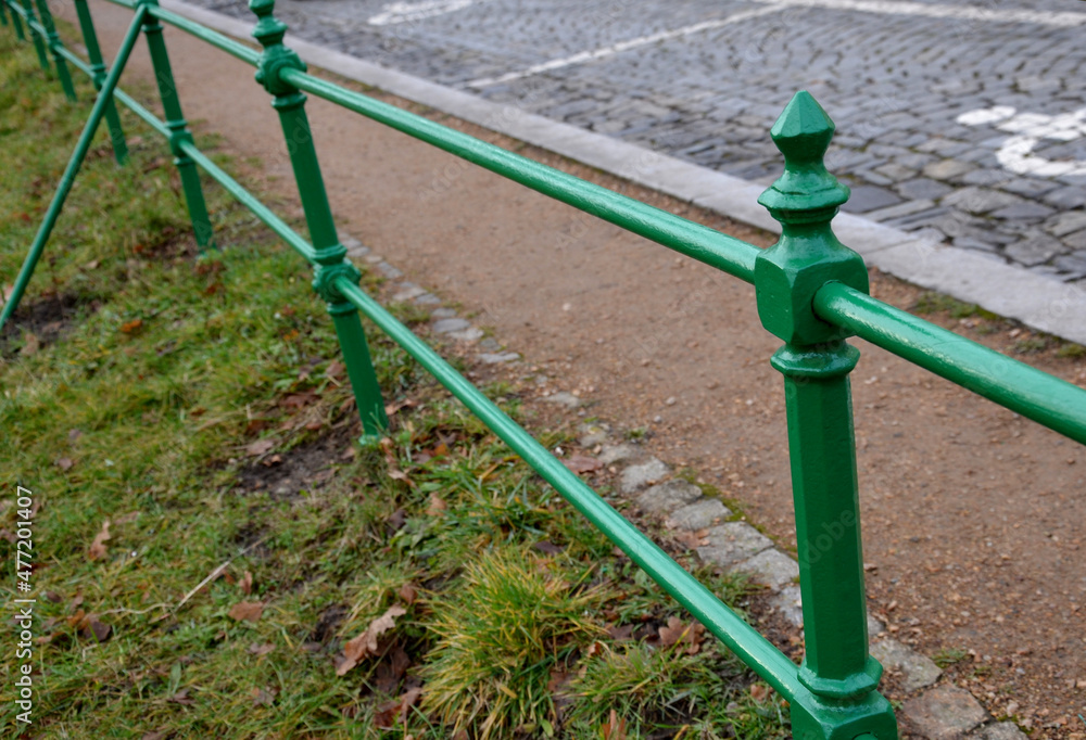green metal railing on the edge of the park by the road made of cobblestones. A metal ornate historic low fence symbolically separates the gravel sidewalk and lawn