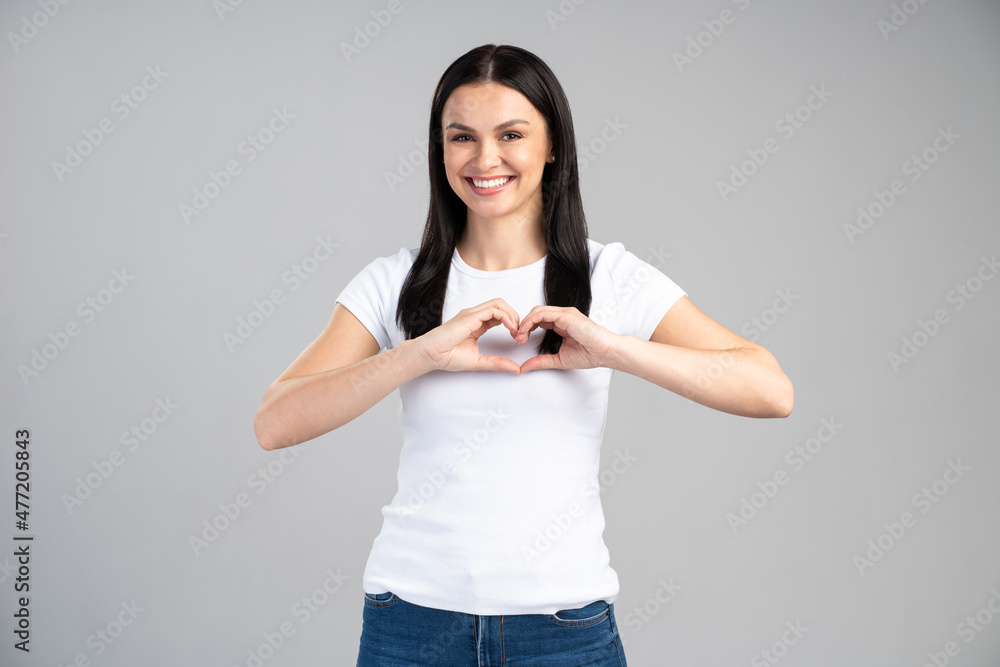 Love you. Portrait of attractive romantic young woman standing and making heart with hands, while smiling playfully. Indoor studio shot isolated on grey background Love you. Portrait of attractive romantic young woman standing and making heart with hands, while smiling playfully. Indoor studio shot isolated on grey background