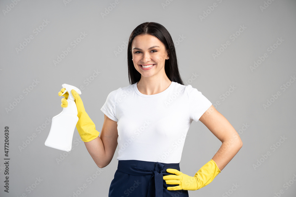 Smiling housemaid wearing rubber protective gloves and holding cleaning spray while being glad to clean everything. Busy cute woman doing spring cleaning at home Smiling housemaid wearing rubber protective gloves and holding cleaning spray while being glad to clean everything. Busy cute woman doing spring cleaning at home