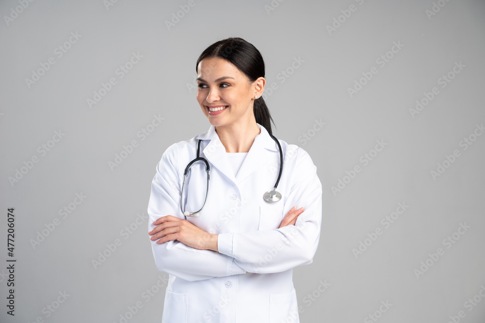 Smiling female doctor in lab coat with arms crossed looking away and posing against grey background. Medicine concept Smiling female doctor in lab coat with arms crossed looking away and posing against grey background. Medicine concept