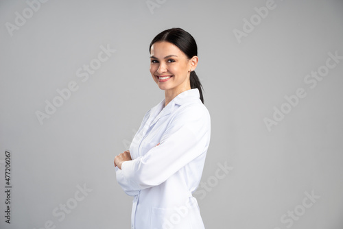 Side view of the smiling female doctor in lab coat with arms crossed looking away and posing against grey background. Medicine concept