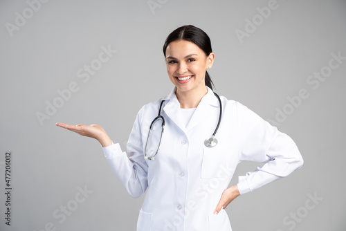 Waist up portrait view of the smiling happy doctor pointing with her palm at the free space on grey background. Doctor and medicine concept