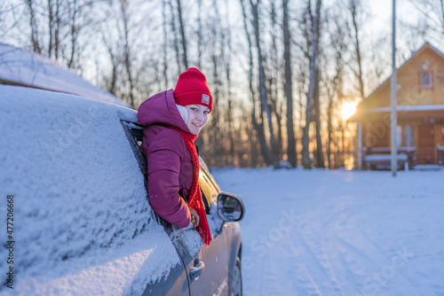 young woman in a red hat peeking out of a snow-covered car on the background of a winter road and the setting sun.