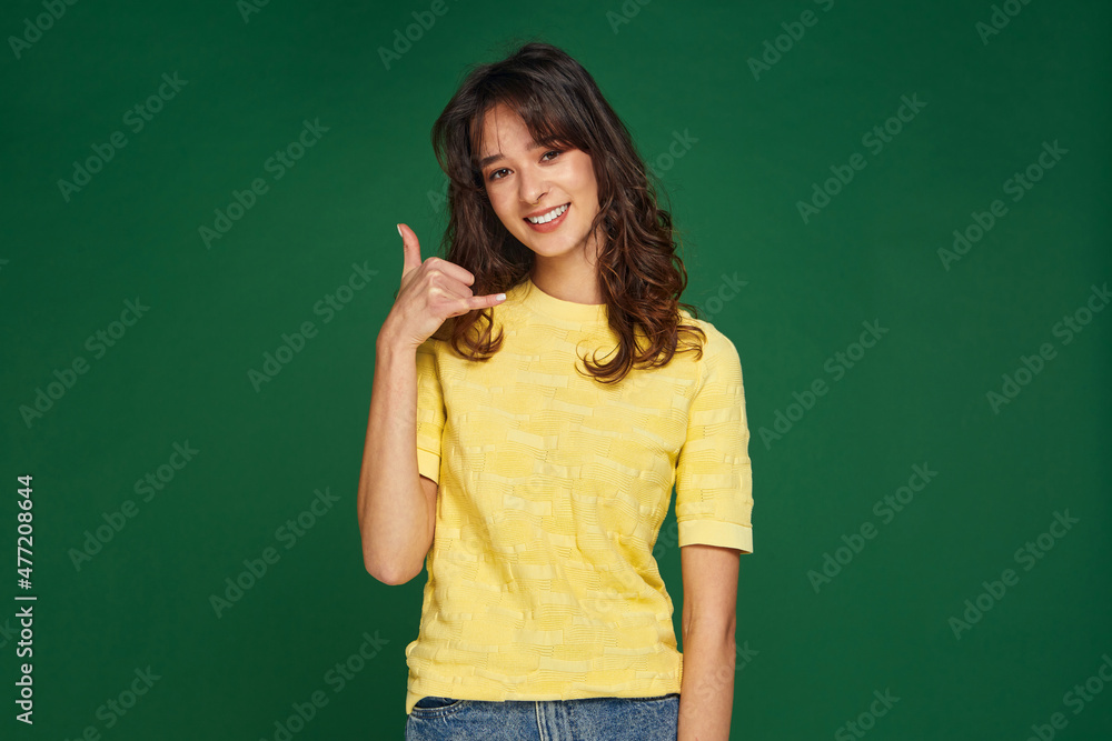 Call me gesture. Friendly modern smiling young girl offering contact communication by phone on green studio background