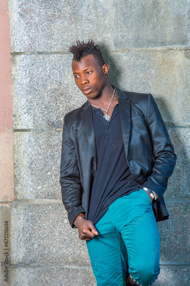 Obraz premium Dressing in a black blazer, green pants, a young black guy with mohawk hair is standing against the wall, looking down into deeply thinking..