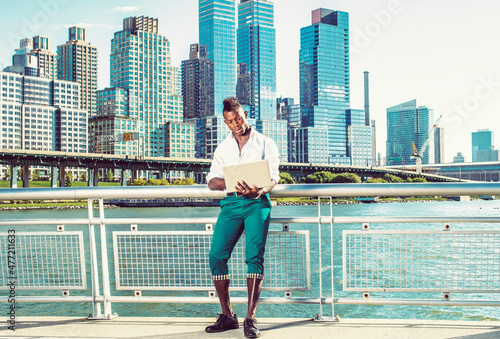 Working Outside. Dressing in a white shirt, green pants, leather shoes, a young black guy with mohawk hair is standing in the front of high buildings, looking down, working on a computer..