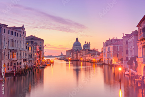 Romantic Venice at dawn, sunrise. Cityscape image of Grand Canal in Venice, with Santa Maria della Salute Basilica reflected in calm sea. Street lights reflected in calm water. © tilialucida
