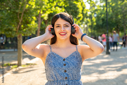 Young woman at park using a dress during summer spring season at a sunny day. Brunette with long black hair using headphones to listen to music